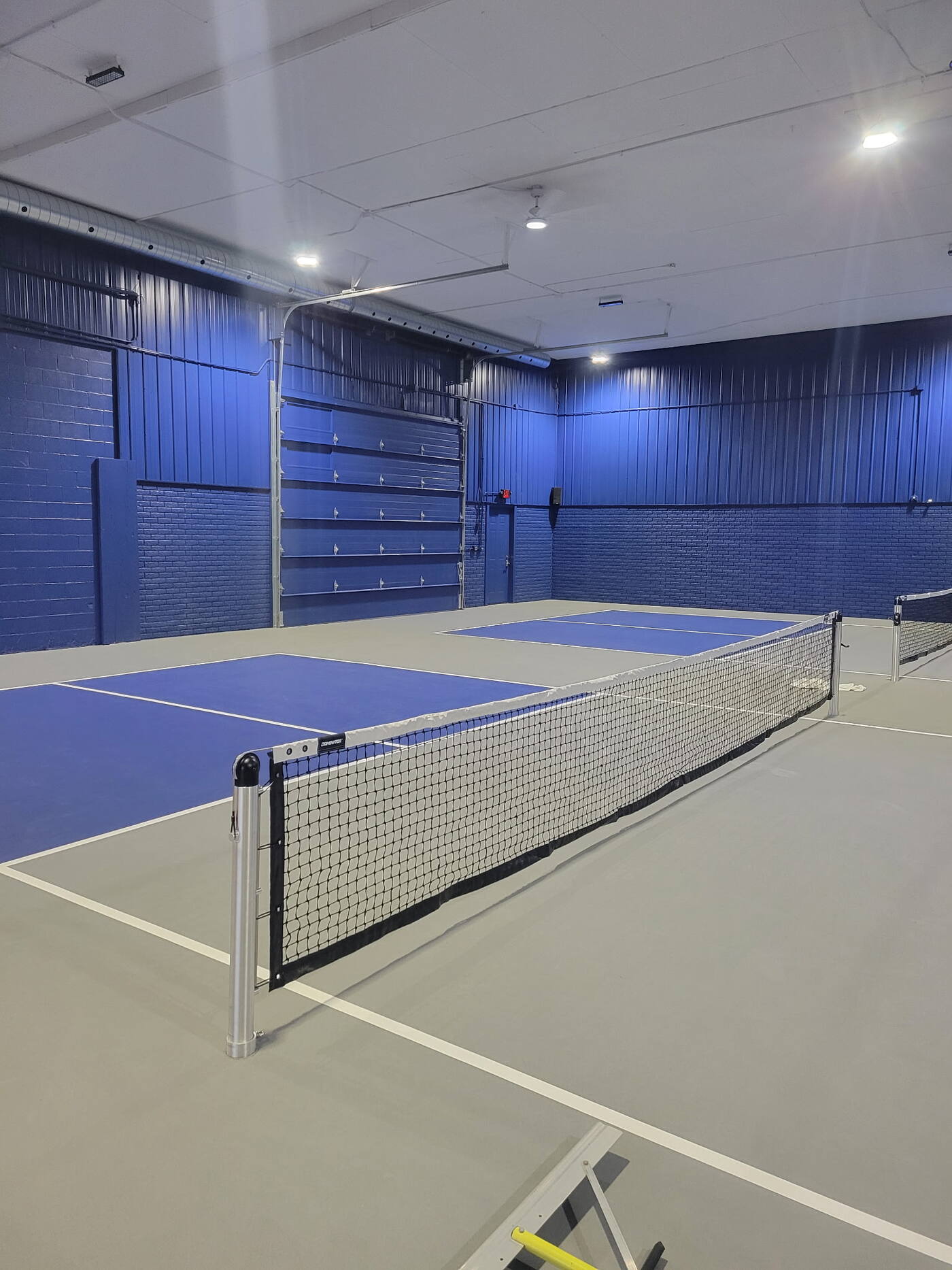 Indoor pickleball facility with vivid blue painted courts, blue accent walls, and gray coated flooring